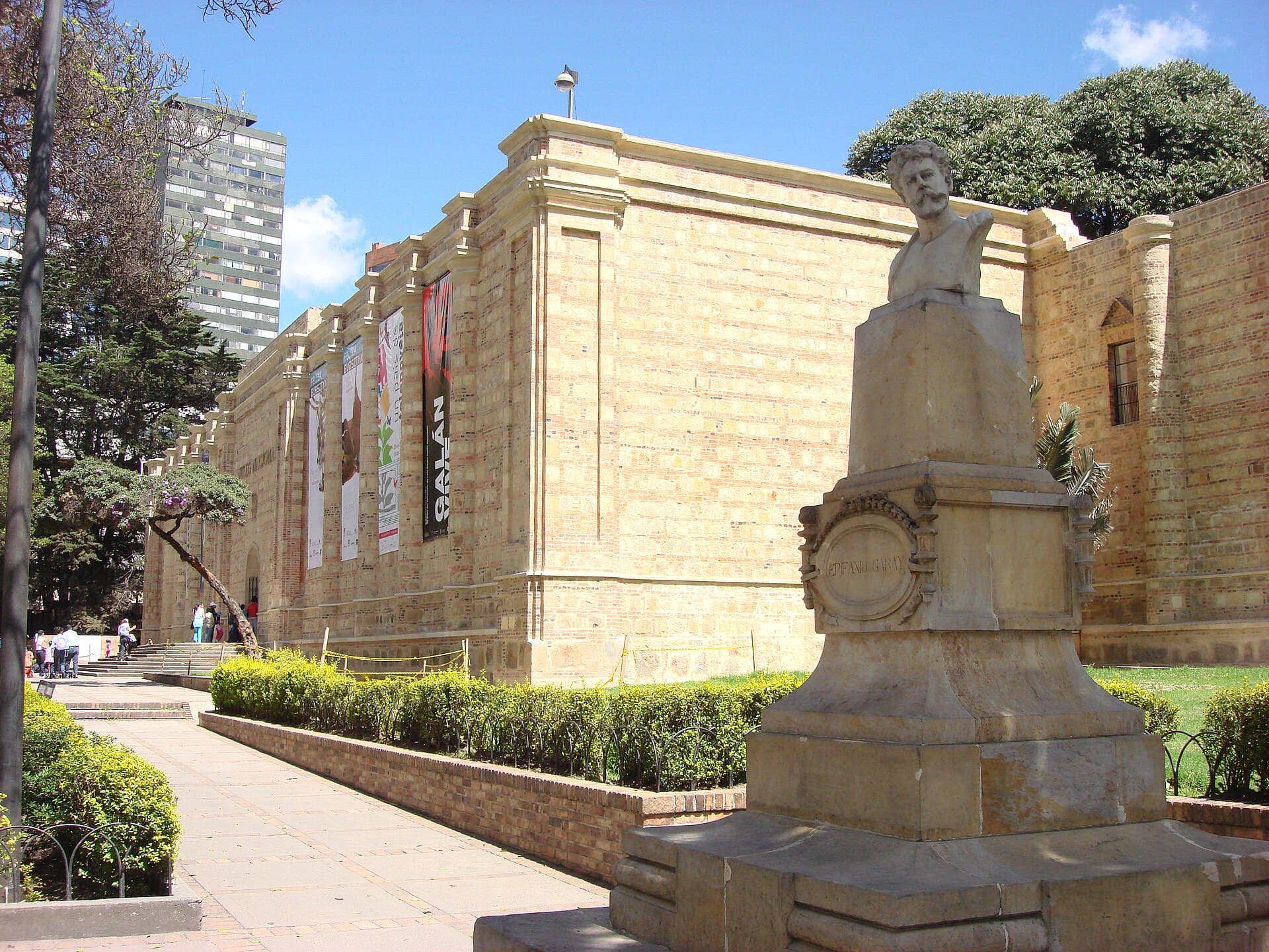 Fachada de piedra del Museo Nacional de Colombia, con pendones de exposiciones y un busto en primer plano.