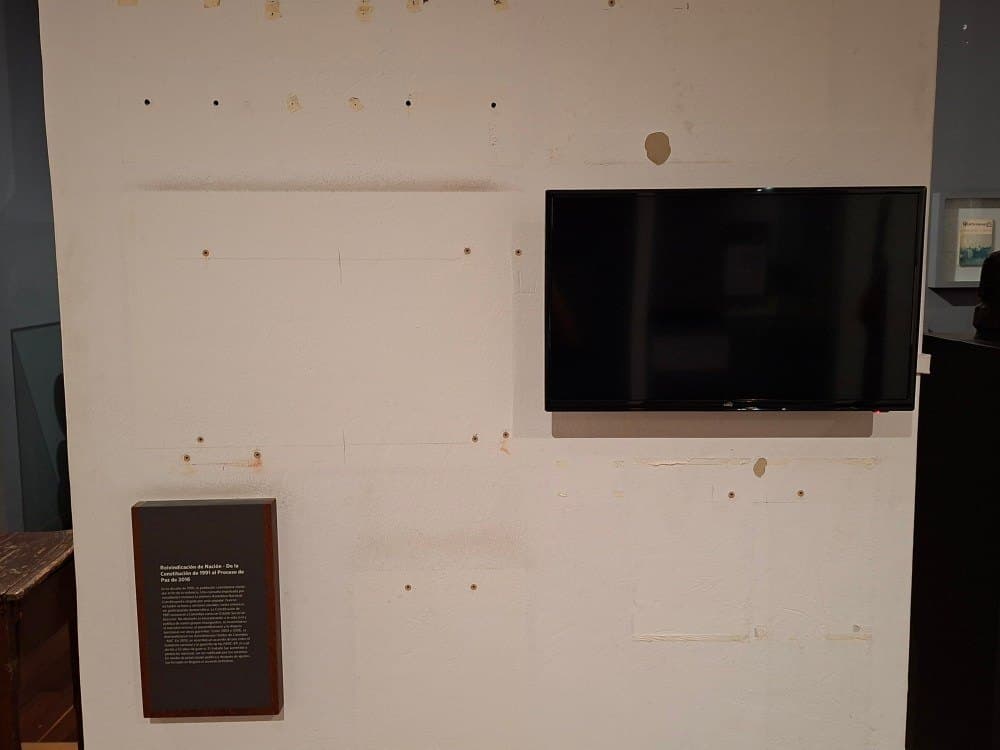 Pared blanca con marcas de soportes, una pantalla apagada y una placa que dice «Reivindicación de Nación».
